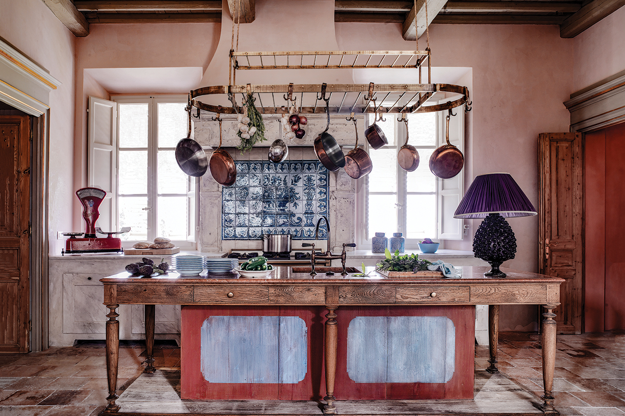 Rustic Tuscan farmhouse kitchen with reclaimed wooden farm table island, plaster walls, hanging cookware, and terracotta tile floors
